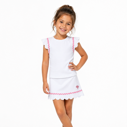 A young girl with her hair in a high ponytail smiles while modeling a white two-piece outfit consisting of a short-sleeve top and a matching skirt with pink scalloped trim and a small floral detail.