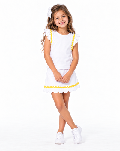 A young girl with long light brown hair wearing a white two-piece outfit with yellow scalloped trim, featuring a short-sleeve top with ruffle details and a matching skirt. She is also wearing a white hair bow and white sneakers, standing against a solid white studio background.