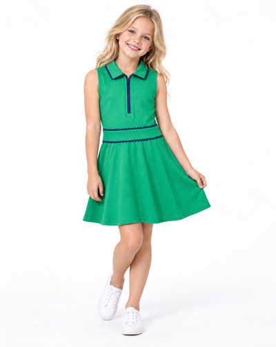 A young girl with blonde hair is modeling a bright green sleeveless polo dress with dark blue trim, paired with white sneakers, against a plain white studio background.