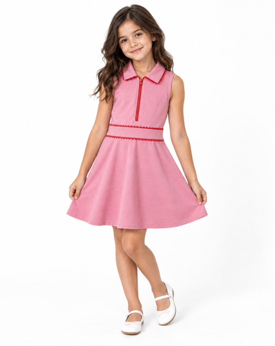 A young girl with long brown hair stands against a white studio background wearing a sleeveless pink dress with red trim and white Mary Jane shoes.