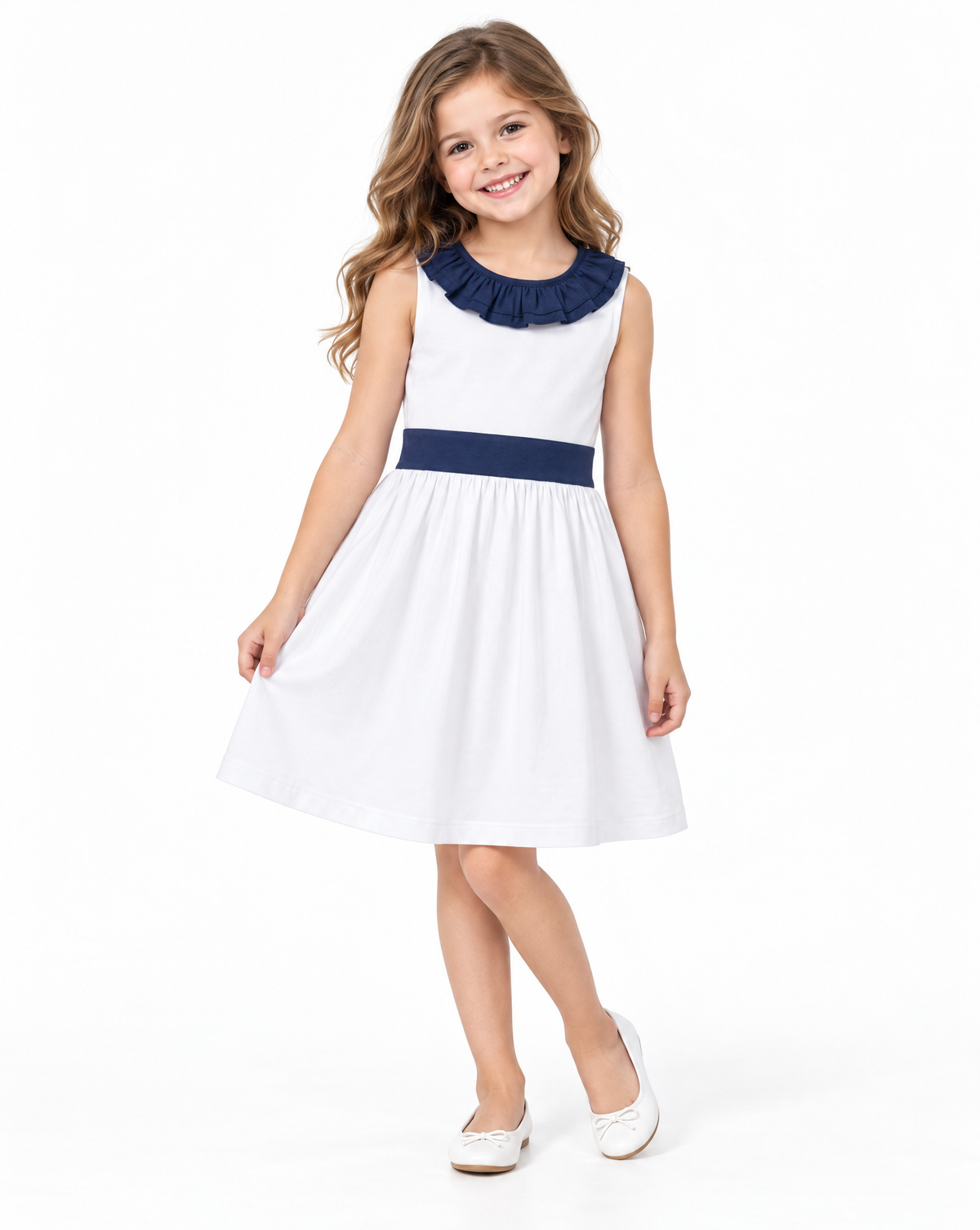 A young girl with wavy light brown hair stands against a white studio background wearing a sleeveless white dress with a navy blue ruffled collar and matching navy waistband, paired with white flats.
