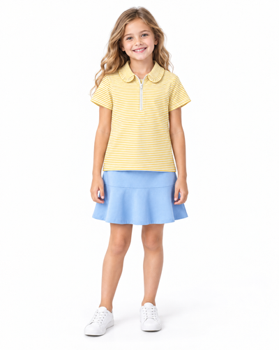 A young girl stands against a white studio background wearing a yellow and white striped short-sleeve polo shirt with a light blue flared skirt and white sneakers.