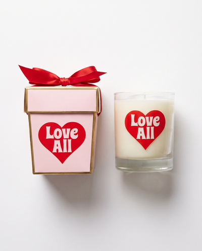 A white candle in a clear glass jar with a red heart label reading 'Love All' sits beside a pink gift box with a red ribbon and matching heart label on a white background.