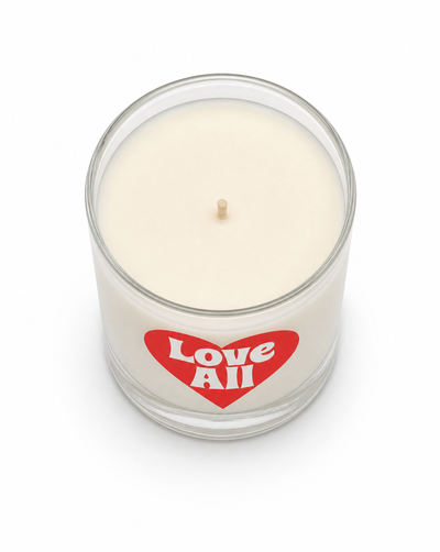 An off-white wax candle in a clear glass jar with a red heart-shaped label that reads 'Love All' in white text, set against a plain white background.