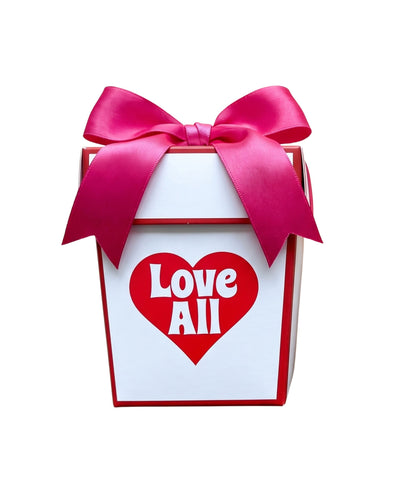 A white gift box with red trim, tied with a large pink satin bow, featuring a red heart on the front with the words 'Love All' in white.