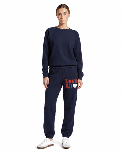 A woman modeling a navy blue sweatshirt and matching jogger pants set. The jogger pants feature the text 'Love All' in red and a white heart on the left thigh.