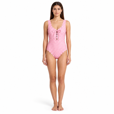 A woman modeling a pink and white gingham one-piece swimsuit with scalloped edges and a lace-up front, standing against a plain white background.