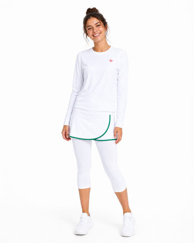 A woman modeling a white long-sleeve athletic top, white capri leggings, and white sneakers against a plain white background. The outfit features a small red logo on the chest and a green curved trim detail along the hem of the top.