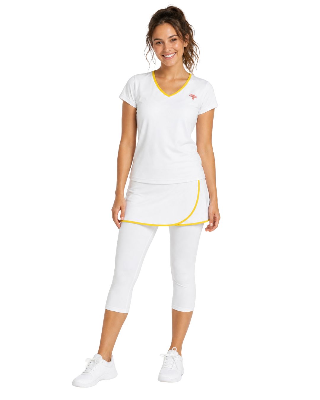 A woman modeling a white athletic set consisting of a V-neck short-sleeve top with yellow trim, a matching mini skirt with yellow piping, white capri leggings, and white sneakers, against a plain white background.