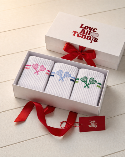 A gift box containing three pairs of white ribbed socks with embroidered tennis-themed designs, shown open on a light wood surface. The box lid has the printed phrase 'Love All Tennis' in red, and the gift box is tied with a red ribbon.