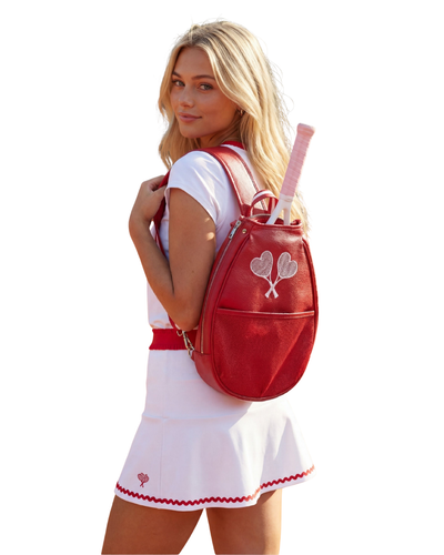 A blonde woman wearing a white tennis outfit and holding a pink tennis racket, shown from the back wearing a red leather tennis backpack with a silver heart design.