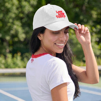 A woman on a tennis court wearing a white baseball cap with red embroidered text that reads 'Love All Tennis' and a white tennis shirt with red trim.