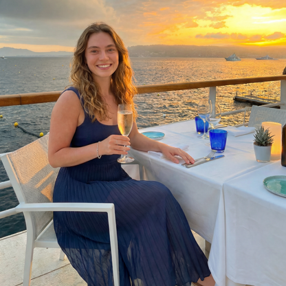 Woman in a blue dress holding a glass of wine at a sunset table by the water.