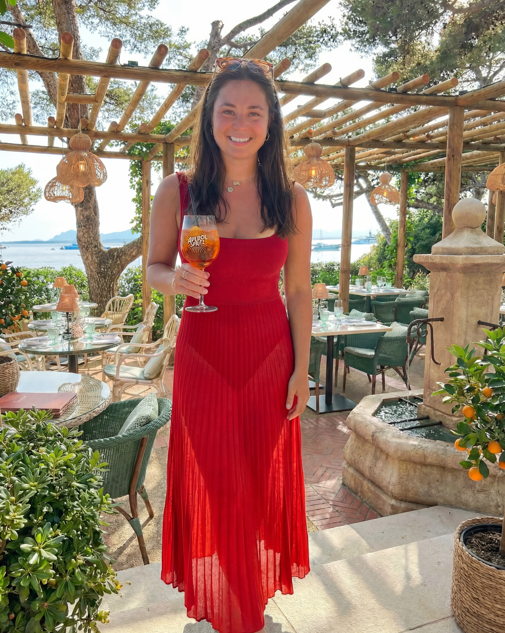 Woman in a red dress holding a glass of orange juice outdoors with tables and chairs in the background.