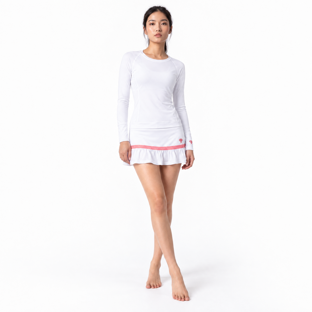 A woman modeling a white long-sleeve athletic top and skirt with a pink ruffled hem and a small red logo on the sleeve, set against a plain white studio background.