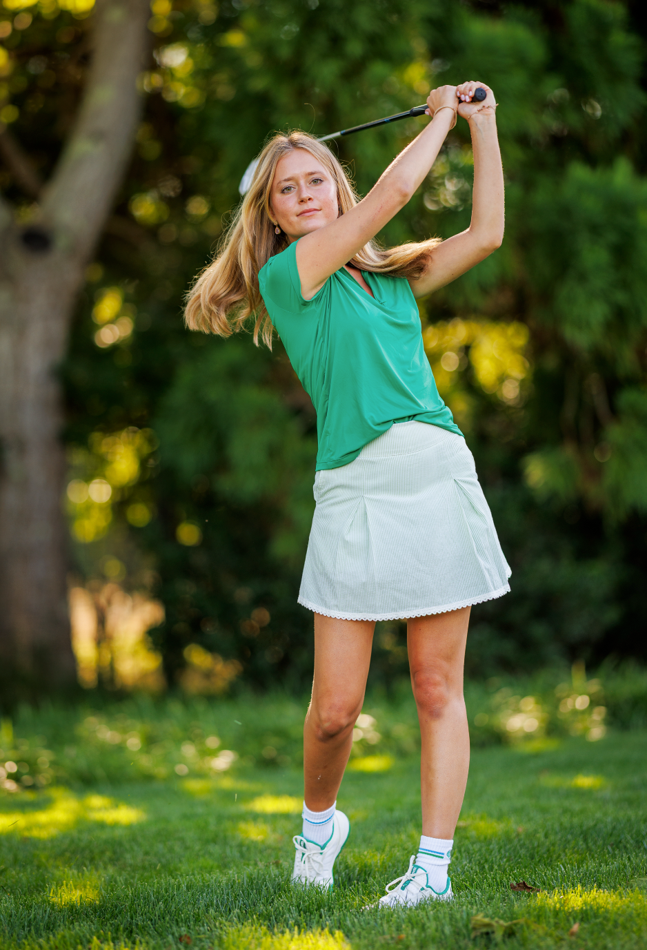 A woman in a green t-shirt and white pleated golf skirt swings a golf club on a sunny grassy golf course.
