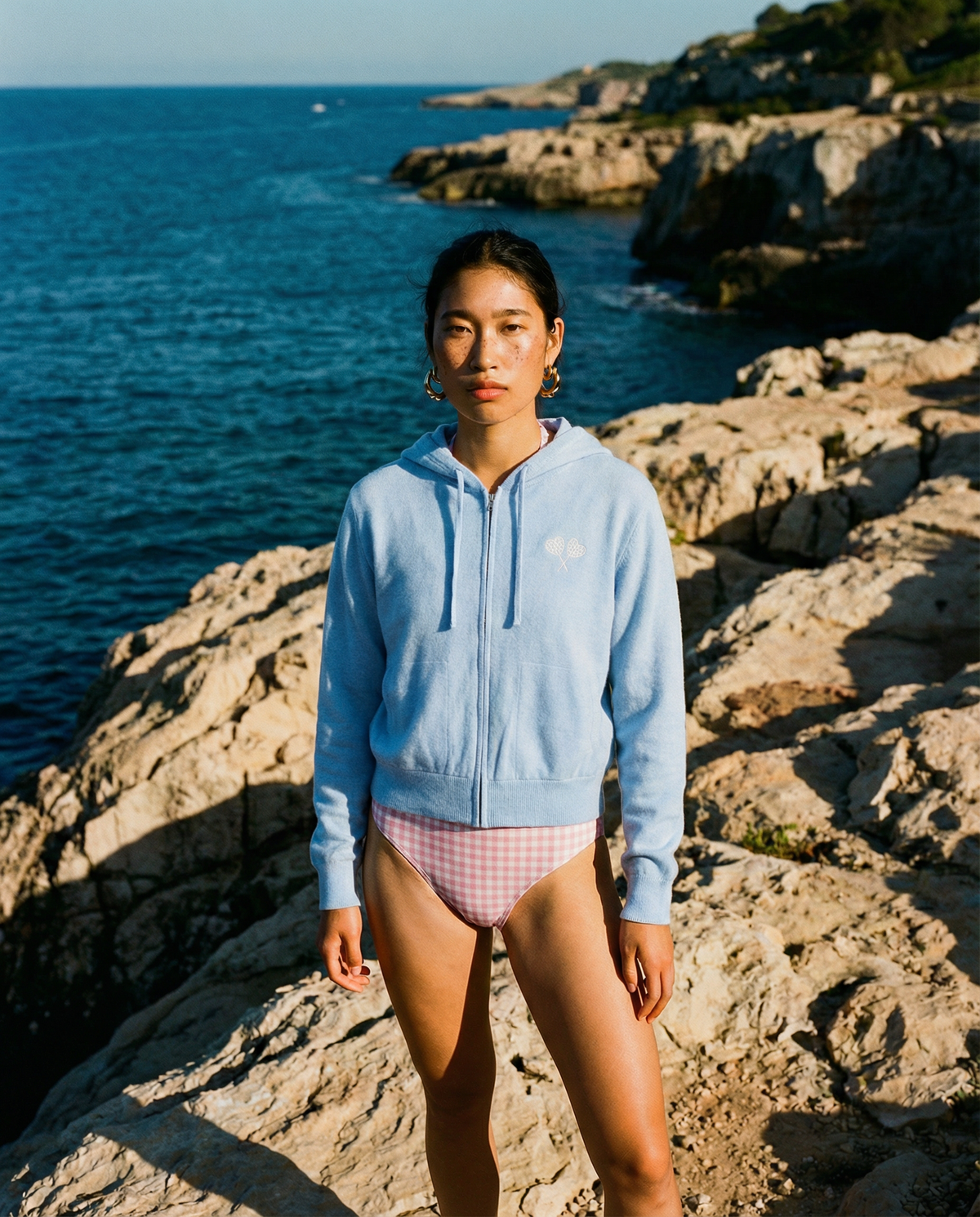 A woman stands on rocky coastal cliffs wearing a light blue zip-up hoodie and pink-and-white gingham bikini bottoms, with the blue ocean behind her.