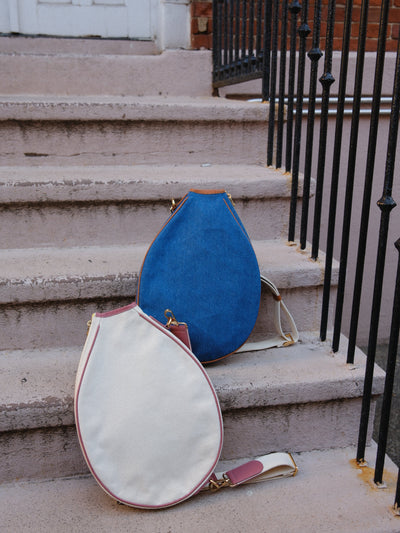 Two teardrop-shaped tennis bags resting on outdoor concrete steps next to a black metal railing. One bag is cream canvas with pink trim, and the other is blue suede with cream trim.