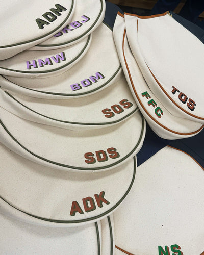 A close-up of several canvas bags stacked together, each with different embroidered letters or initials in contrasting colors along the front and border piping.