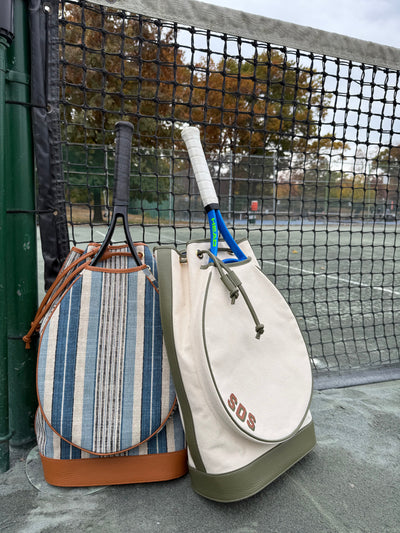 Two tennis bags and two tennis rackets leaning against a tennis court net, with a tennis court and trees in the background.