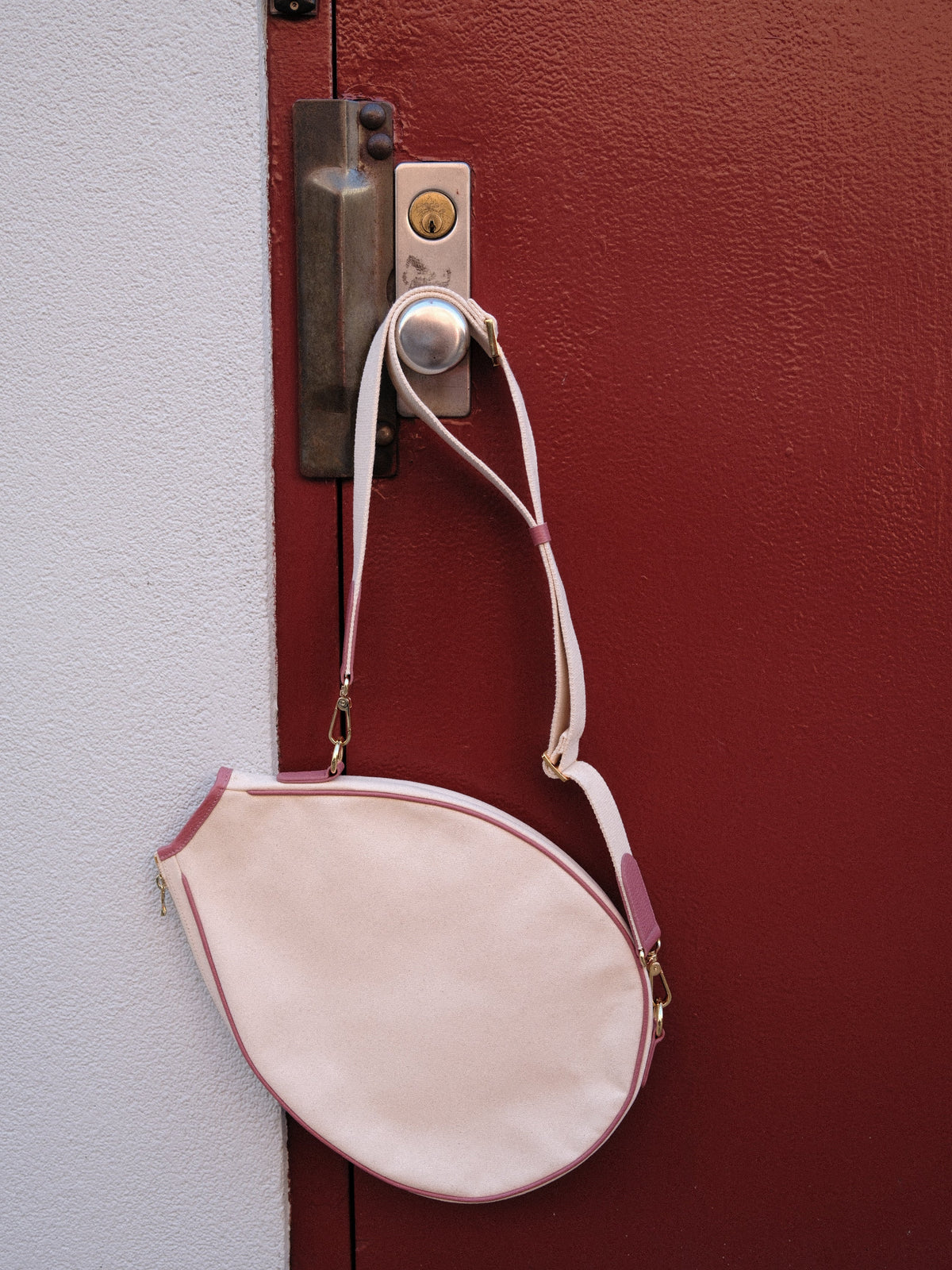 A light pink teardrop-shaped shoulder bag with a thin pink strap, hanging from a silver doorbell on a textured red door next to a white wall.