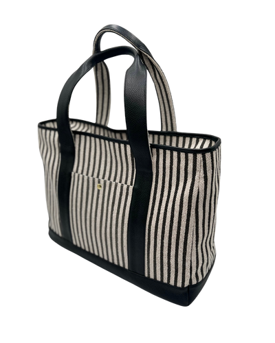 A striped tote bag with black leather handles, trim, and base, featuring a vertical cream and black stripe pattern and a small gold logo on the front pocket.