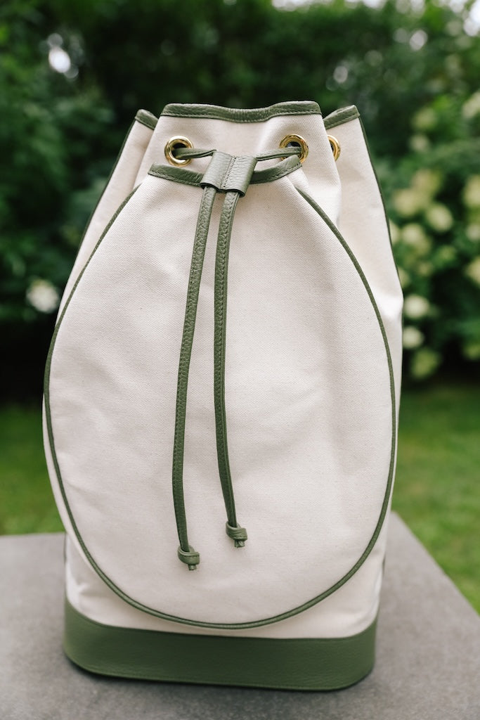 An off-white canvas drawstring backpack with olive green leather trim, drawstring ties, and a green base, set against a blurred green garden background.