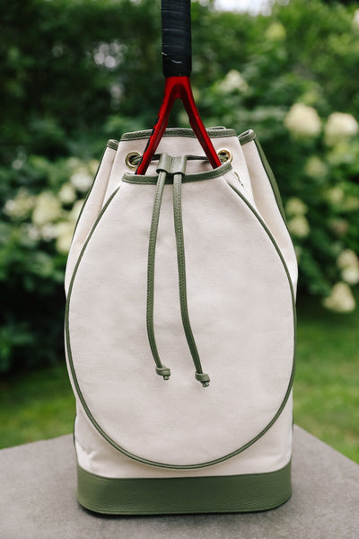 A canvas drawstring bucket bag in off-white and olive green, featuring a large oval front pocket and a black-and-red hanging strap, set against a blurred garden background.