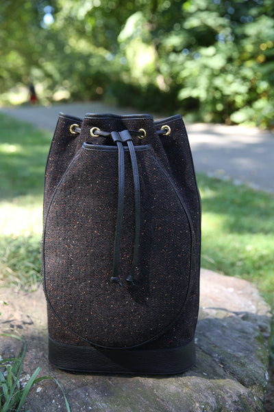 A dark brown textured bucket bag with black drawstrings and gold-tone eyelets, resting on a stone in an outdoor park setting with green grass and trees in the background.