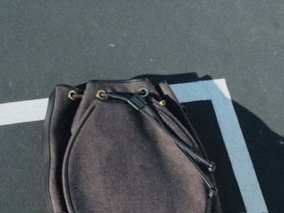 A dark grey denim drawstring bucket bag with gold-tone metal eyelets, resting on asphalt next to a white painted line.