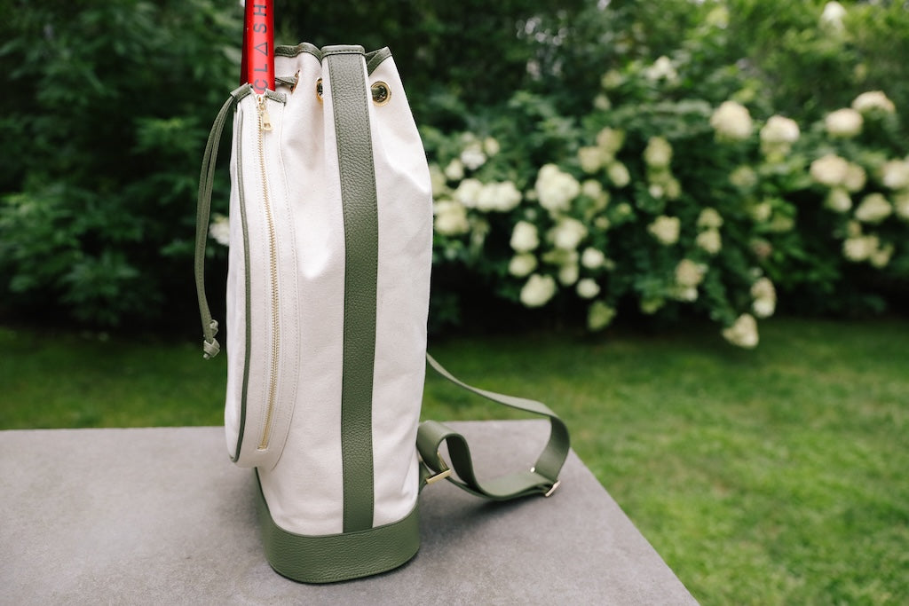 A white canvas golf bag with olive green leather trim and a green shoulder strap, standing on a stone surface with a blurred garden background of green grass and white flowers.