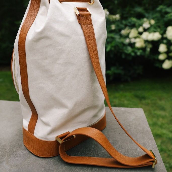 A canvas bucket bag with tan leather trim and a matching shoulder strap, resting on a stone surface with a blurred garden background.