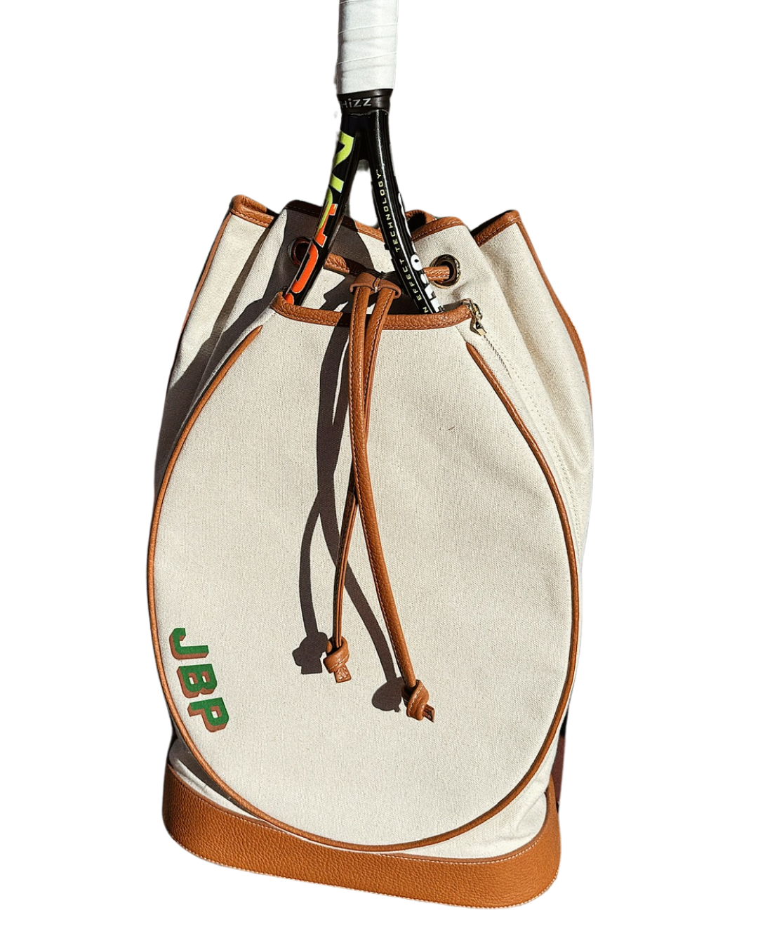 An off-white canvas drawstring bag with brown leather trim and a green 'JBP' logo, shown with a tennis racket tucked inside on a white background.