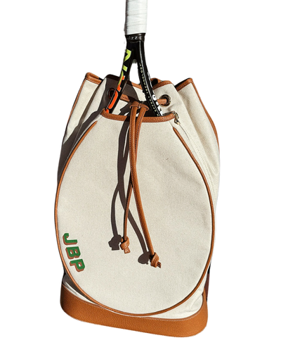 An off-white canvas drawstring bag with brown leather trim and a green 'JBP' logo, shown with a tennis racket tucked inside on a white background.