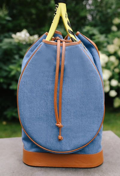 A blue denim-like drawstring backpack with tan leather trim and handles, shown outdoors on a stone surface with green foliage blurred in the background.