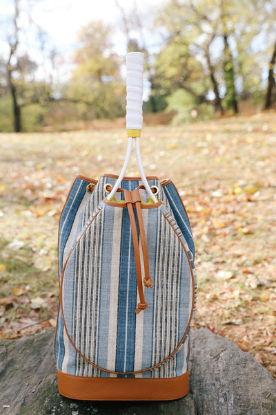 A striped bucket bag with blue, beige, and white vertical stripes, tan leather trim, and a white wrapped handle, sitting on a stone in an outdoor park setting with fallen leaves.