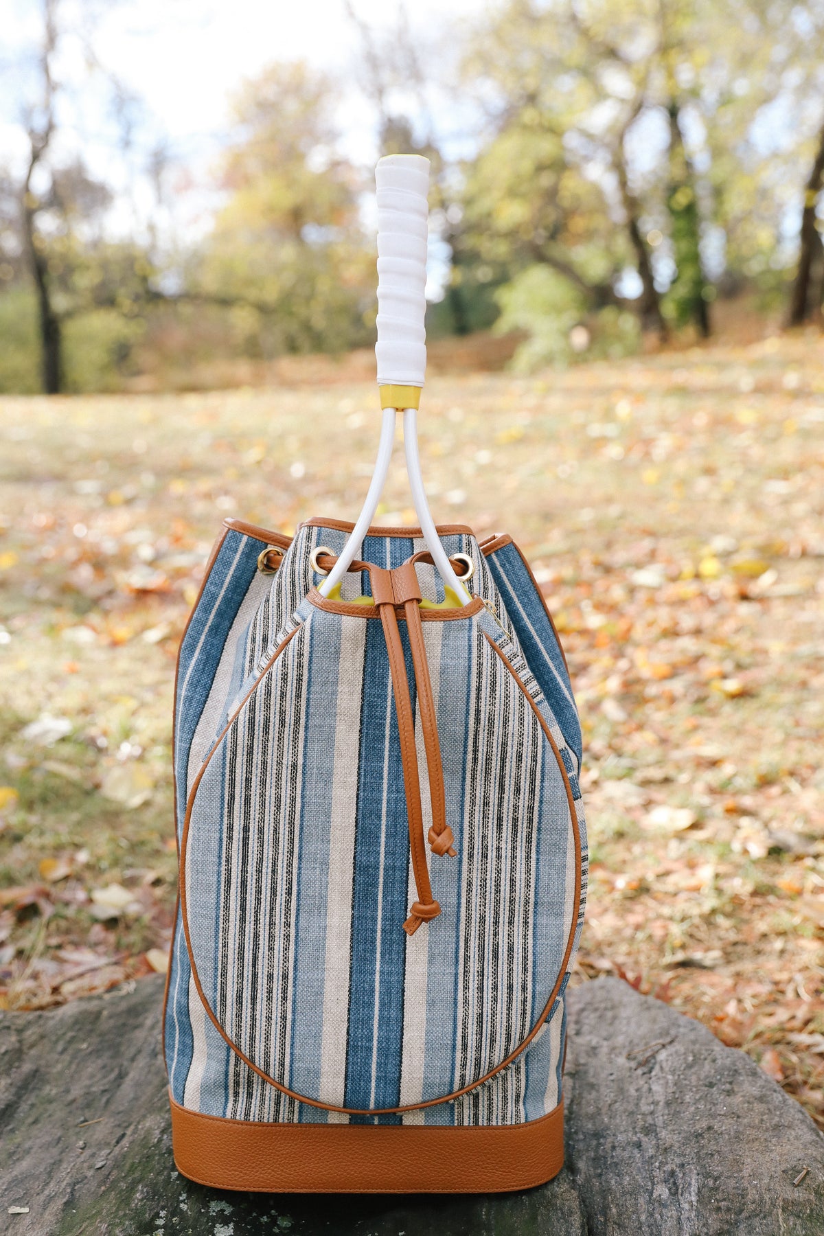 A striped bucket bag with blue, beige, and white vertical stripes, tan leather trim, and a white wrapped handle, sitting on a stone in an outdoor park setting with fallen leaves.