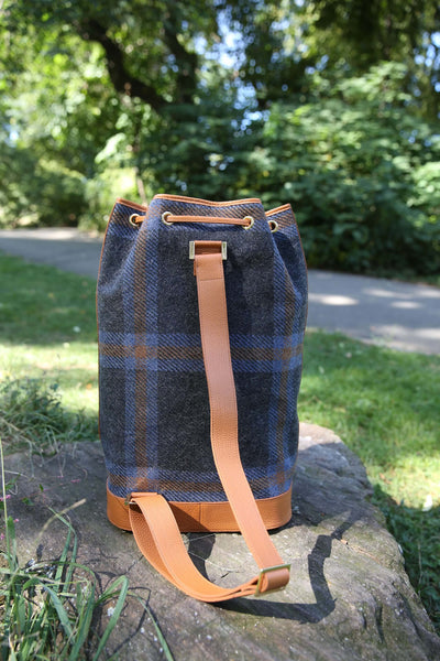 A plaid bucket bag made of dark blue and brown wool, with tan leather trim and strap, sitting on a stone in a sunny park setting.