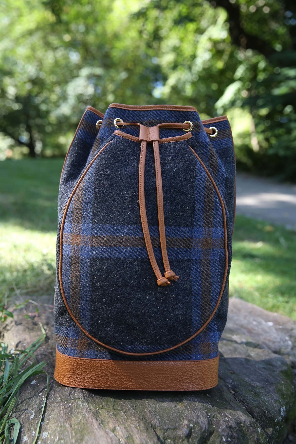 A blue and brown plaid wool bucket bag with tan leather trim and drawstring closure, resting on a stone outdoors with a blurred green garden background.