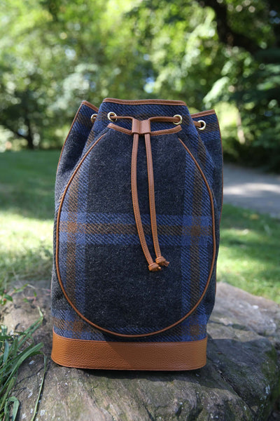 A blue and brown plaid wool bucket bag with tan leather trim and drawstring closure, resting on a stone outdoors with a blurred green garden background.