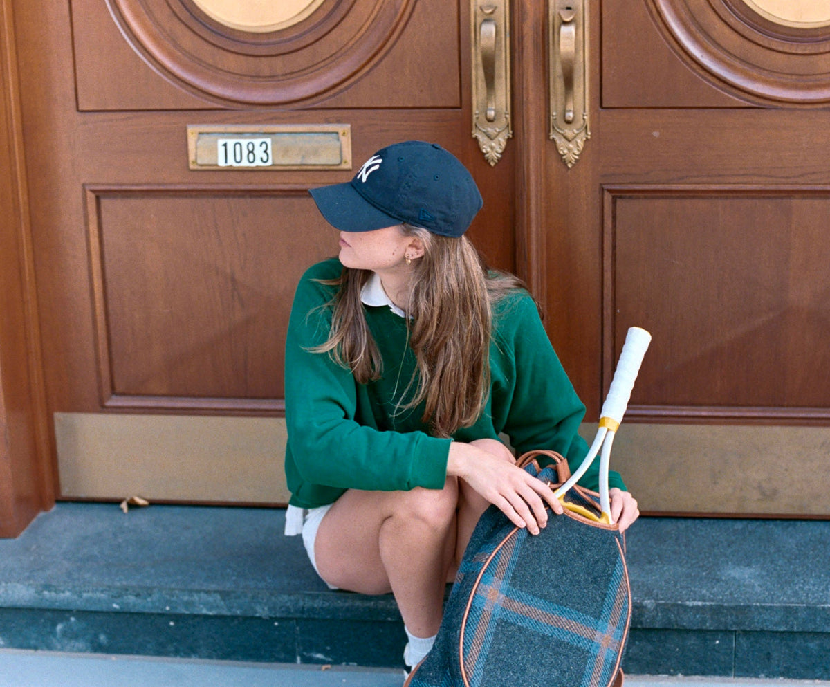 A young woman sits on the steps in front of a large brown wooden door, wearing a dark green sweatshirt, white shorts, a navy baseball cap, and holding a vintage tennis racket and a plaid handbag.
